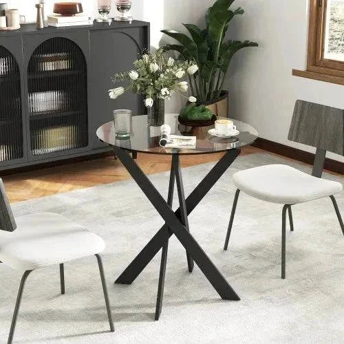 Modern 70cm round tempered glass table with black metal crossed legs and two white chairs in a minimalist dining room setting